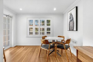 A dining area with a round table and four chairs, a window with blinds, and a framed artwork on the wall. - Pilgrims Apartments (Griffith)