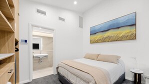 A bedroom with a bed, a bedside table, a nightstand, a wall-mounted TV, a bathroom with a sink, and a painting on the wall. - Pilgrims Apartments (Griffith)