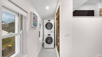Un lavadero con dos lavadoras apiladas, una ventana con vistas al exterior y una puerta que da a otra habitación.