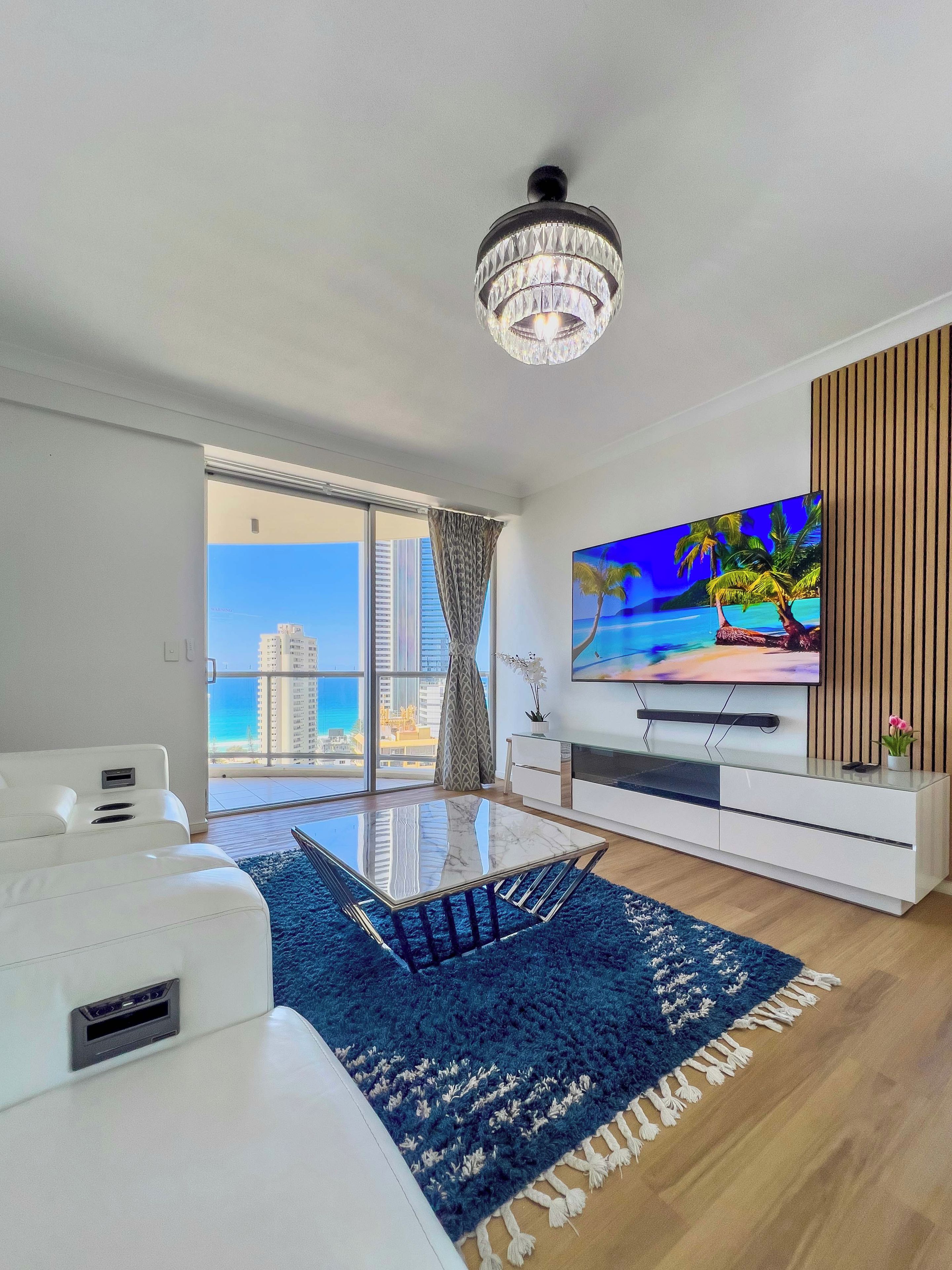 A modern living room with a large TV, a glass coffee table, and a view of the cityscape.