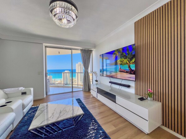 Ein modernes Wohnzimmer mit großem Fernseher, einem weißen Sofa, einem Marmortisch und Blick auf den Ozean.