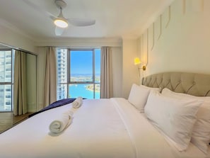 Quarto de hotel com uma cama grande, ventoinha no teto, janela vista para o mar e luminária fixada na parede.