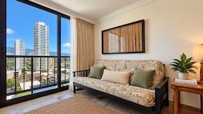 A living room with a sofa, a window with a view of buildings, a framed artwork, and a small table with a plant. - Waikiki Tropical Retreat  Walk to Beach + Lanai (Honolulu)