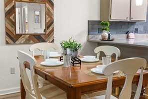 A dining area with a wooden table set for four, surrounded by white chairs. There are potted plants on the table, and a kitchen area is visible in the background. - Foothills Getaway 2BR Home (Branson)