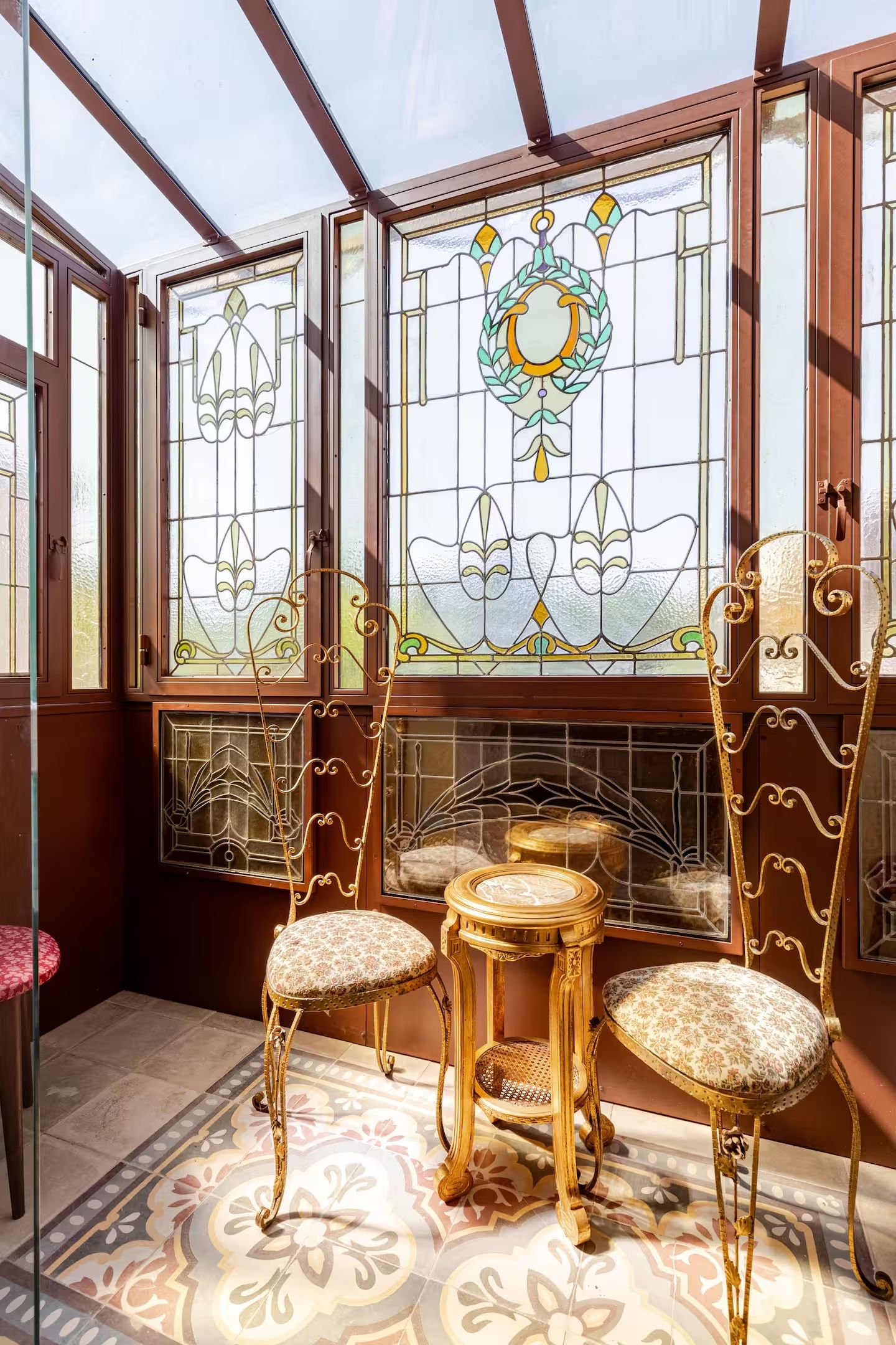 Un invernadero con puertas de cristal ornamentadas, una chimenea y dos sillones con tapicería floral.