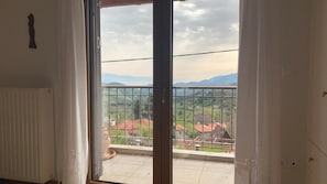 A room with a view of a balcony overlooking a landscape with houses and mountains.