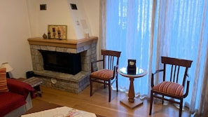 Una sala de estar con chimenea de piedra, dos sillones de madera, una mesita redonda y vistas a través de cortinas traslúcidas.