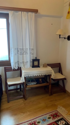 A room with a wooden bench, a table with a lace tablecloth, a wall-mounted lamp, and a window with white curtains.