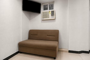 Un sofá pequeño y marrón en un rincón de una habitación con paredes blancas, una ventana con persianas y un aire acondicionado instalado.