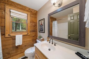 Une salle de bain aux murs en bois, une fenêtre donnant sur les arbres, un lavabo blanc et un grand miroir.