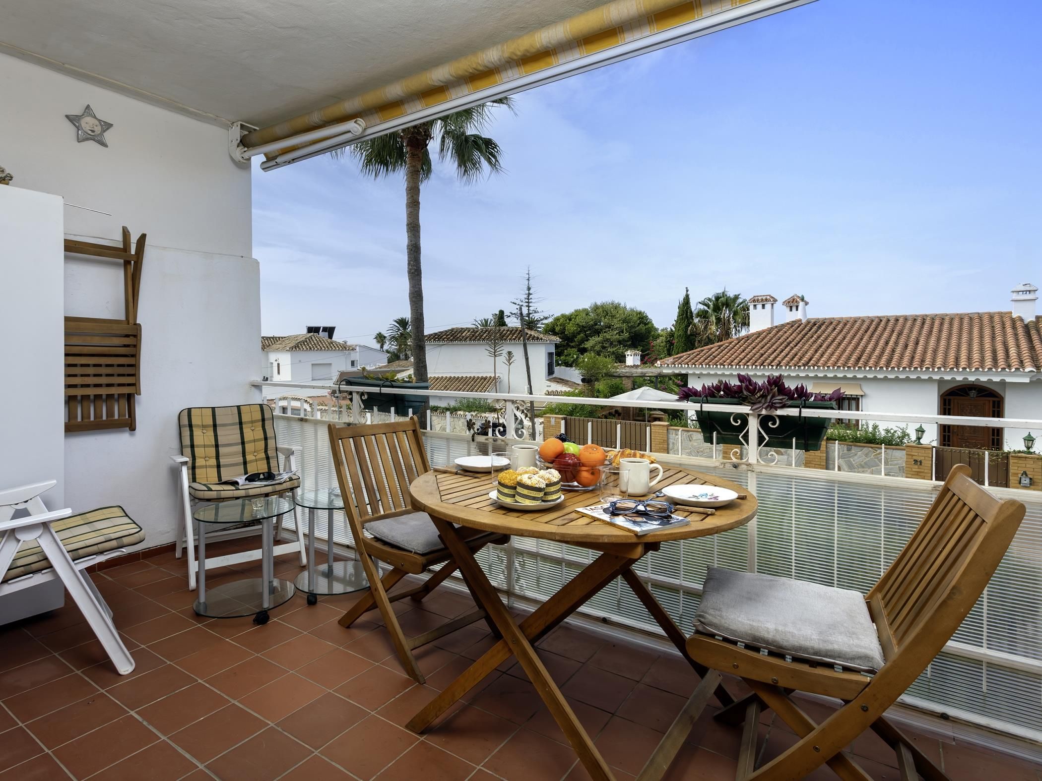 Une terrasse avec une table dressée pour deux personnes, des chaises, un fauteuil avec un coussin et une fenêtre donnant sur un quartier résidentiel avec des palmiers et des maisons.