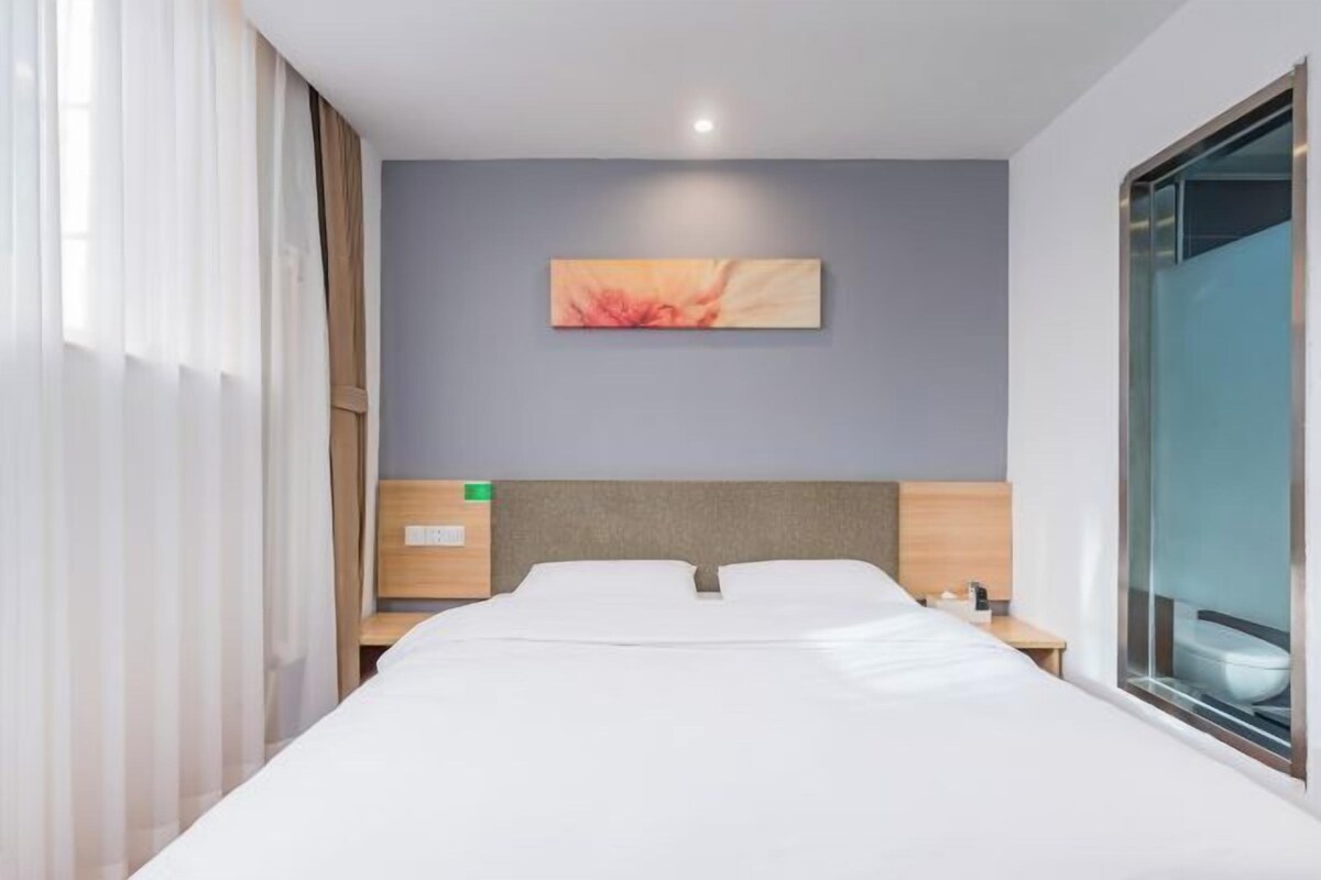 A hotel room with a bed, a nightstand, a wall-mounted painting, and a window with curtains.