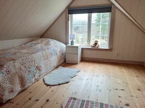Ein Schlafzimmer mit schrägen Wänden, einem Bett mit floralem Überwurf, einem Nachttisch mit Lampe, einem Fenster mit Teddybär und einem Teppich auf dem Holzboden.