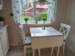 Ein kleiner Essbereich mit Tisch und Stühlen, ein Fenster mit Vorhängen und Blick auf ein rotes Gebäude.