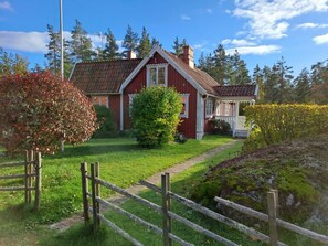 Ein rotes Haus mit weißem Wintergarten, umgeben von einem Holzzaun, Bäumen und einem klaren Himmel.