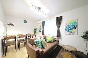 A living room with a brown sofa, wooden chairs, a small round table, a floor lamp, and a large window with curtains. - Homeward Shinkawa D (Sapporo)