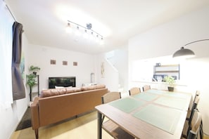 A modern living room with a brown leather sofa, a wooden dining table with chairs, a flat-screen TV, and a staircase leading to an upper level. - Homeward Shinkawa D (Sapporo)