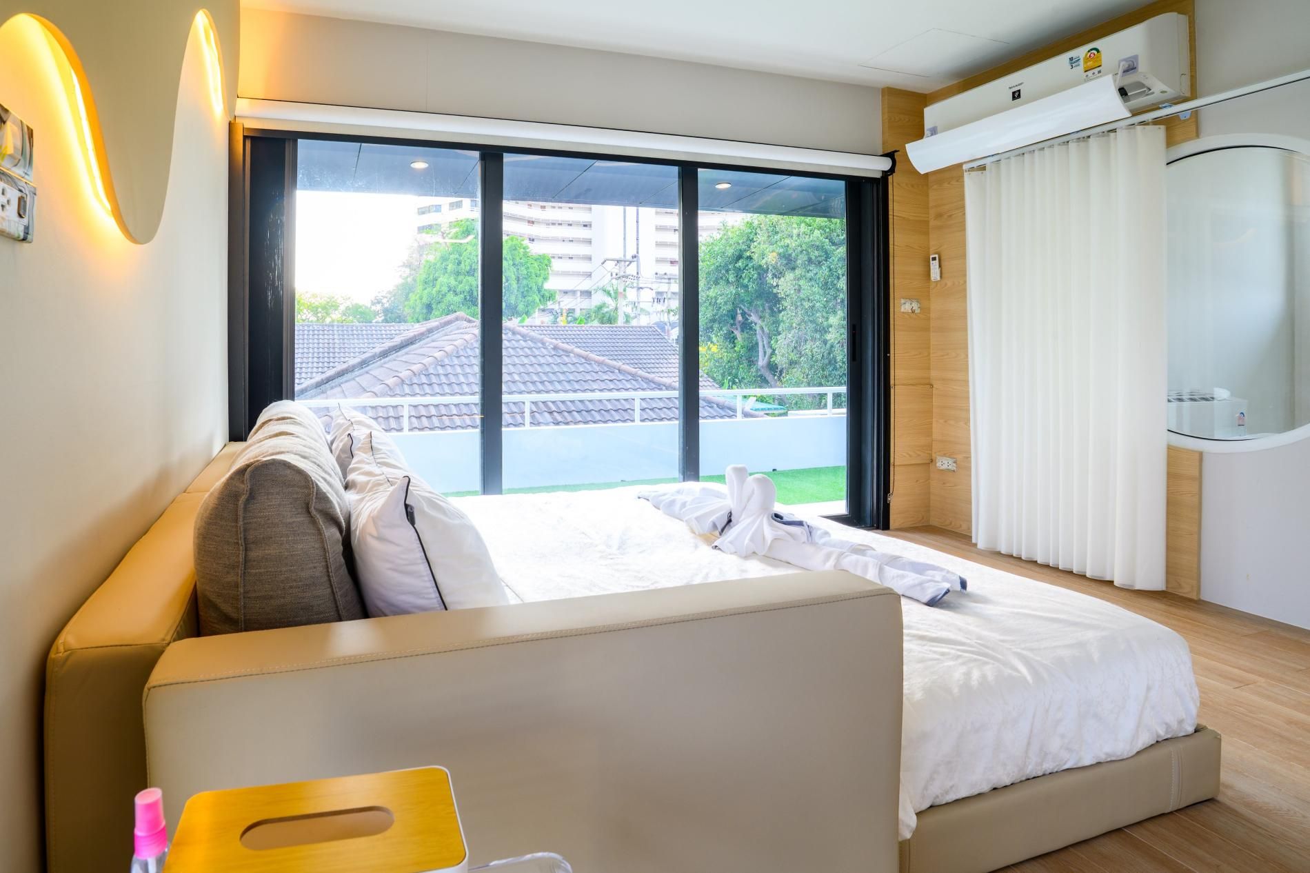 A modern hotel room with a large bed, a wooden headboard, a window with a view of buildings, and a ceiling-mounted air conditioning unit.