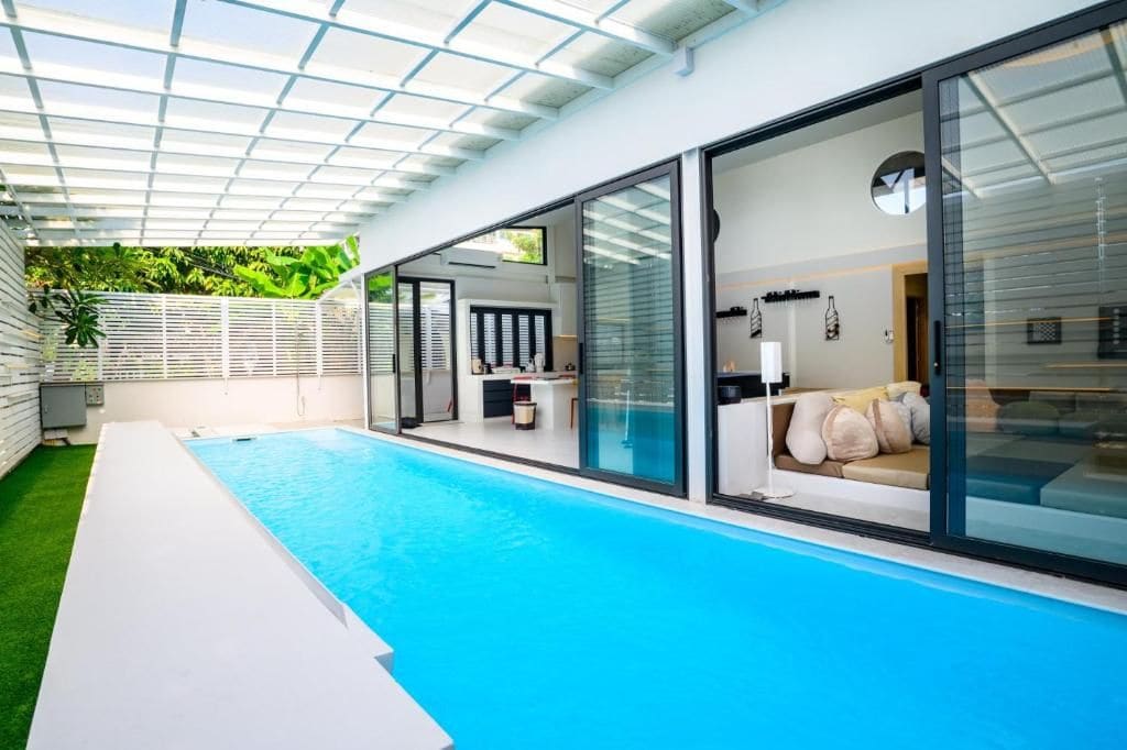 Una piscina interior moderna con paredes de cristal, fachada blanca y zona de césped.