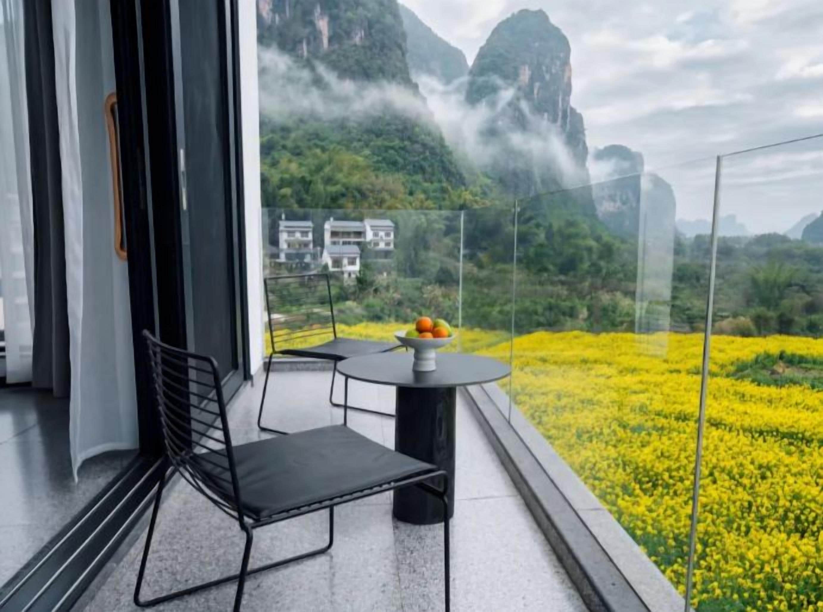 A balcony with a table and chairs, overlooking a field of yellow flowers and a mountainous landscape.