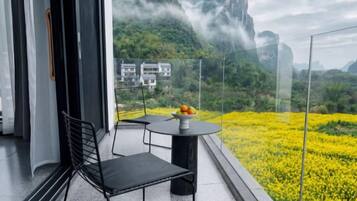 A balcony with a table and chairs, overlooking a field of yellow flowers and a mountainous landscape.