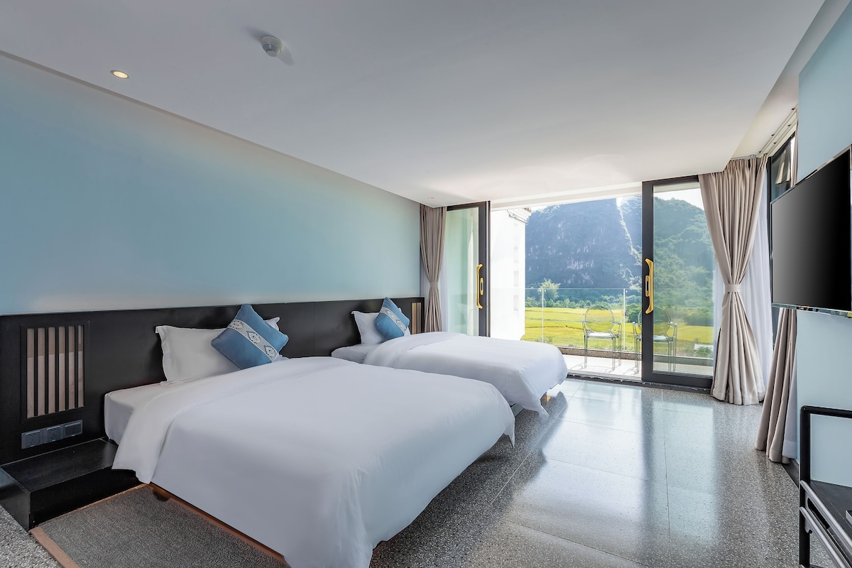 A hotel room with two beds, a large window with a mountain view, and a flat-screen TV.