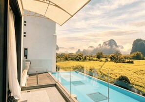 A modern outdoor pool with clear glass panels, a white building with a balcony, and a view of a mountainous landscape.