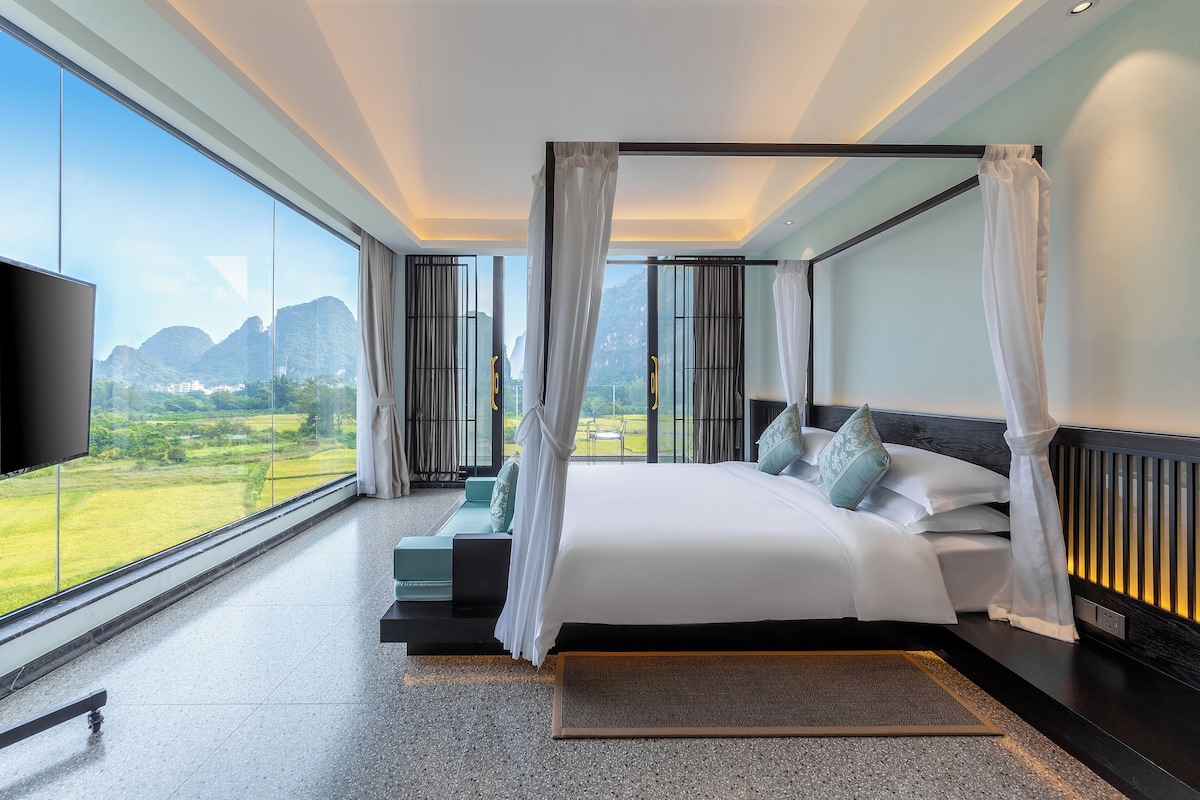 A modern hotel room with a large bed, a TV, and a view of a mountainous landscape.