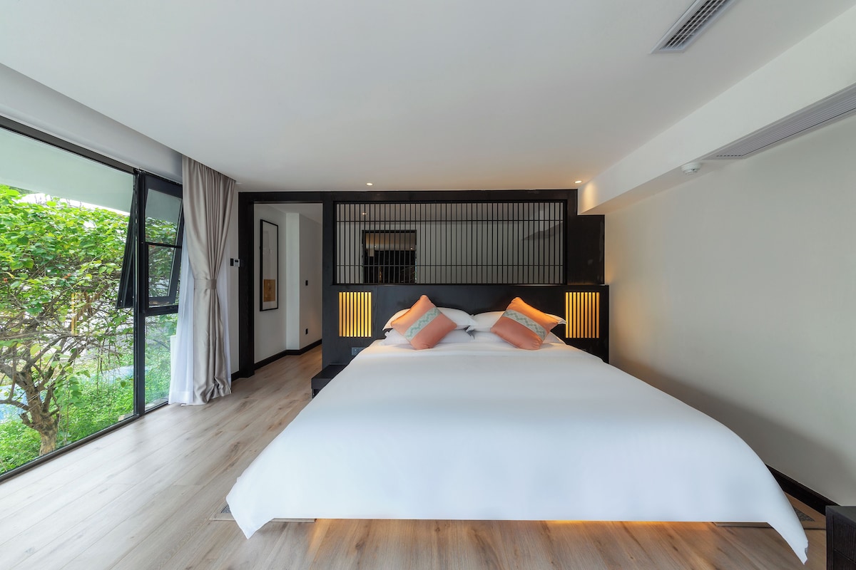 A modern bedroom with a large bed, a view of greenery outside, and a large window.