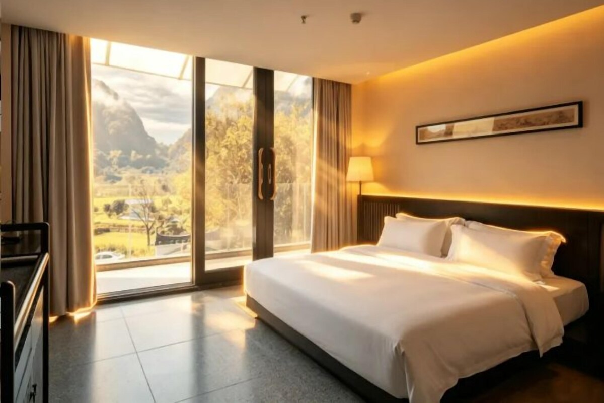 A hotel room with a large bed, a desk, and a view of a mountainous landscape through a glass door.