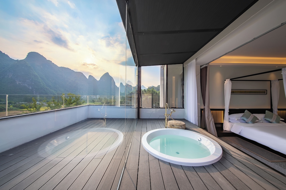 A modern hotel room with a large glass window, a private balcony, a hot tub, and a bed with white bedding.