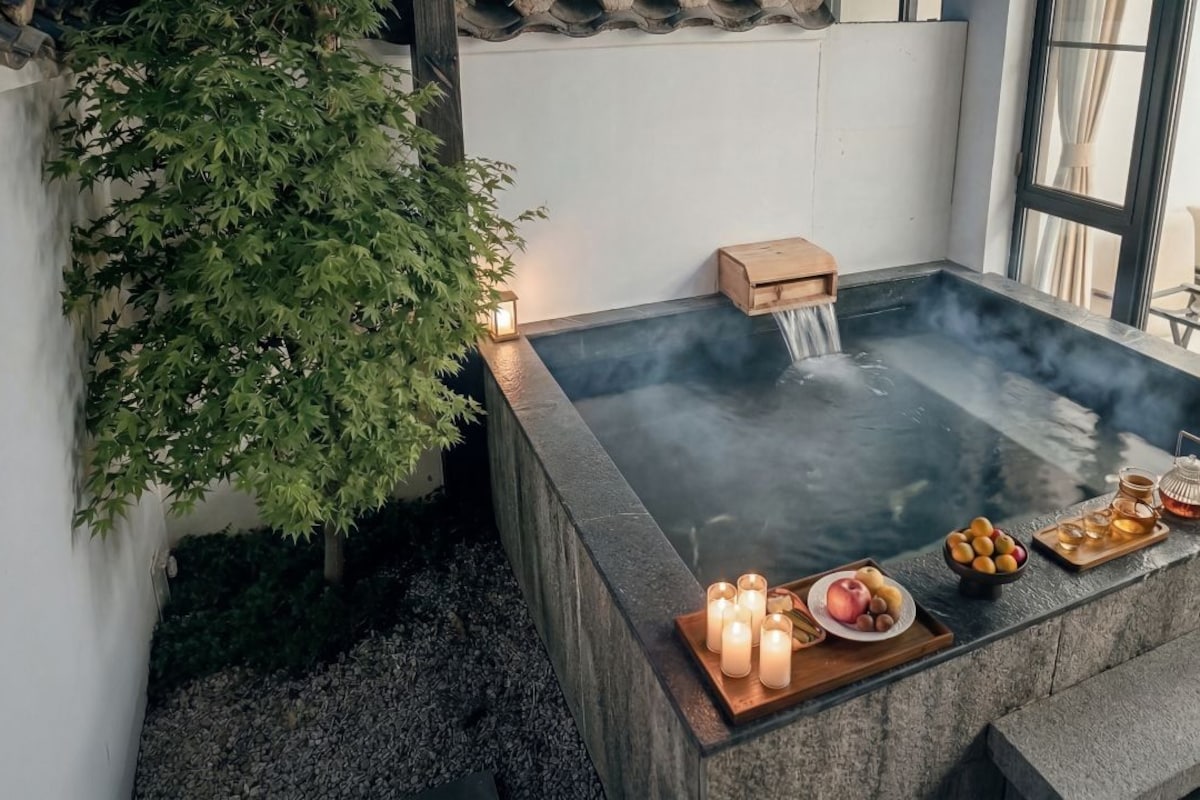 A hot spring with a waterfall feature, surrounded by greenery and a wooden bench.