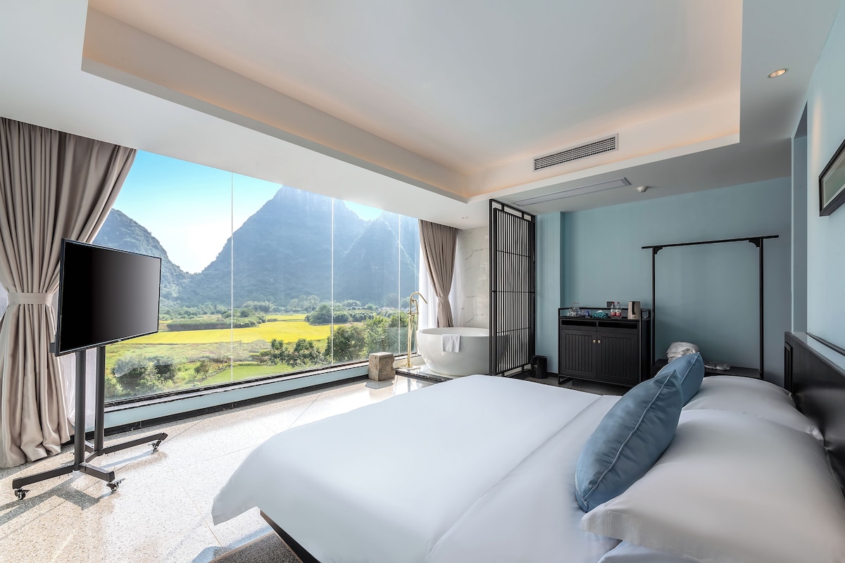 A hotel room with a large bed, a flat-screen TV, a bathtub, and a view of mountains.