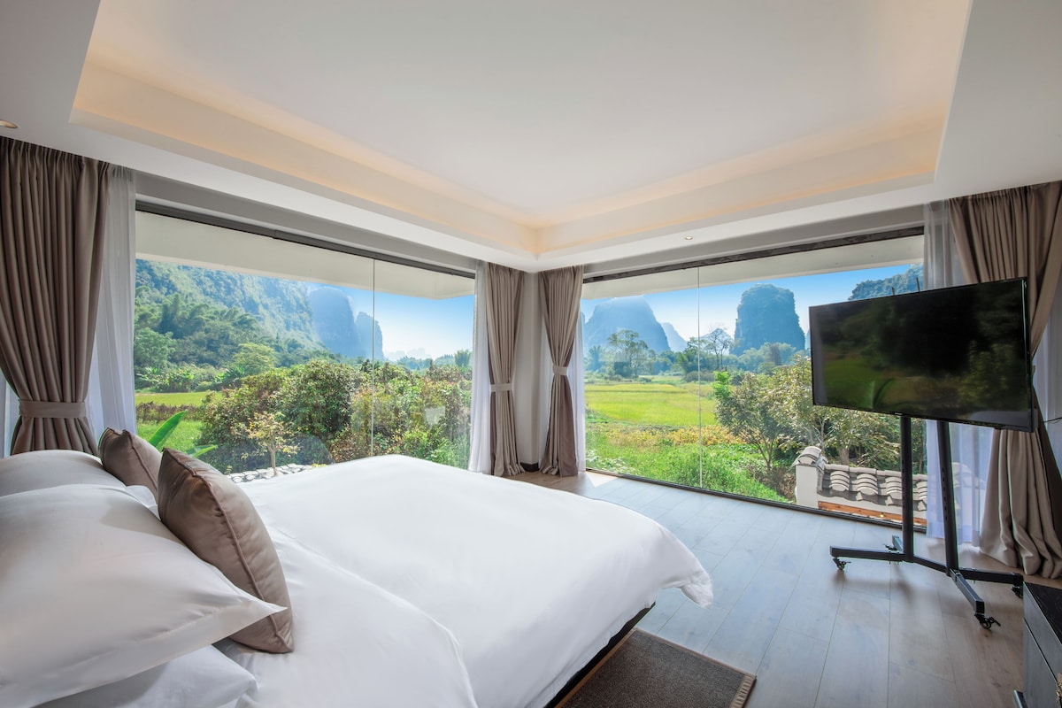 A bedroom with a large bed, a flat-screen TV, and a view of a green landscape through large windows.