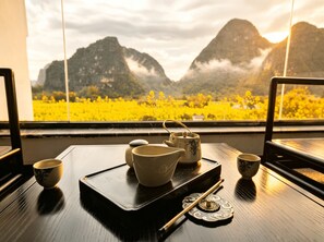 A table with tea set and chopsticks, a tray, and a window with a view of mountains and yellow flowers.