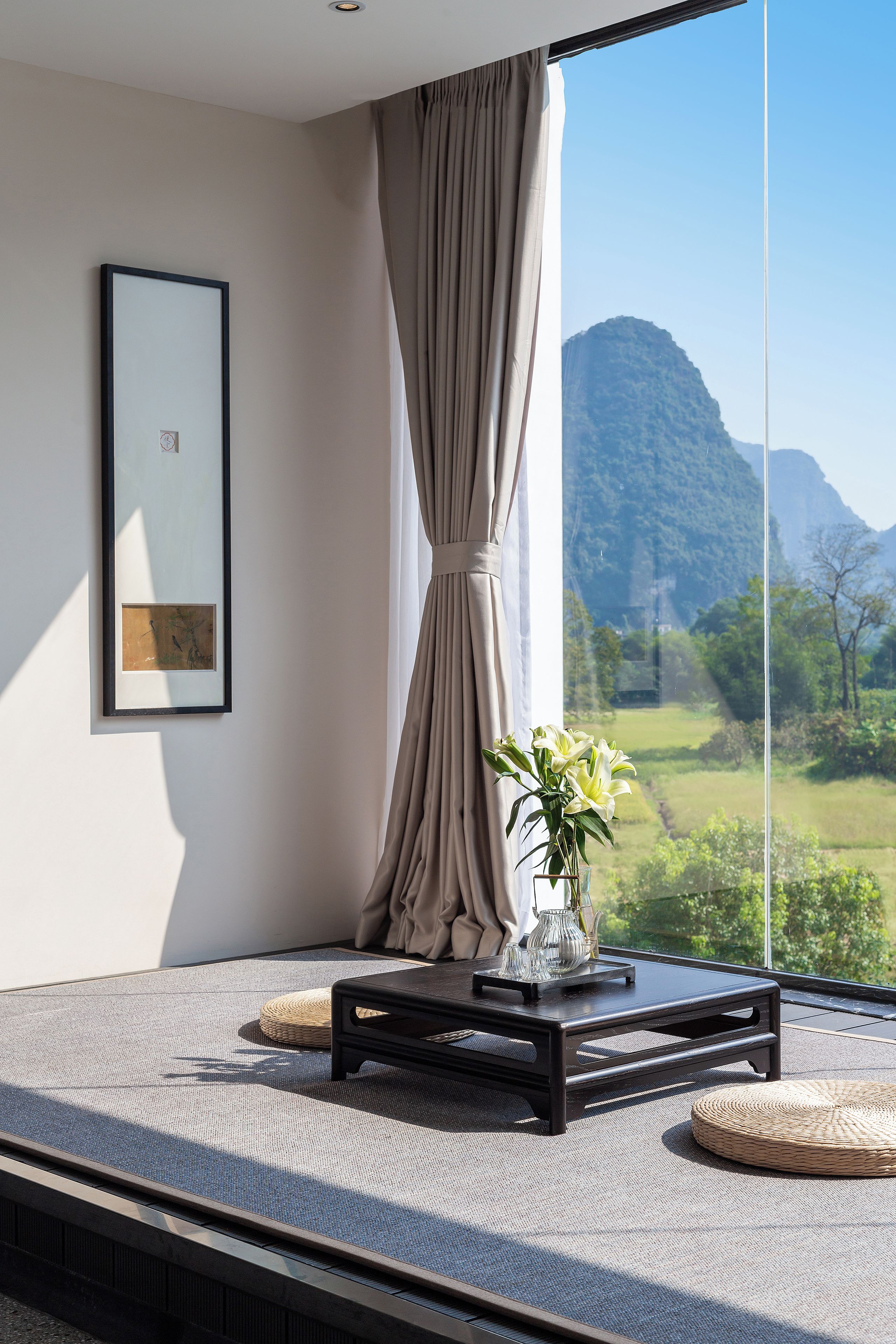 A room with a large window, a framed picture on the wall, a small table with a vase of flowers, and a woven floor mat.