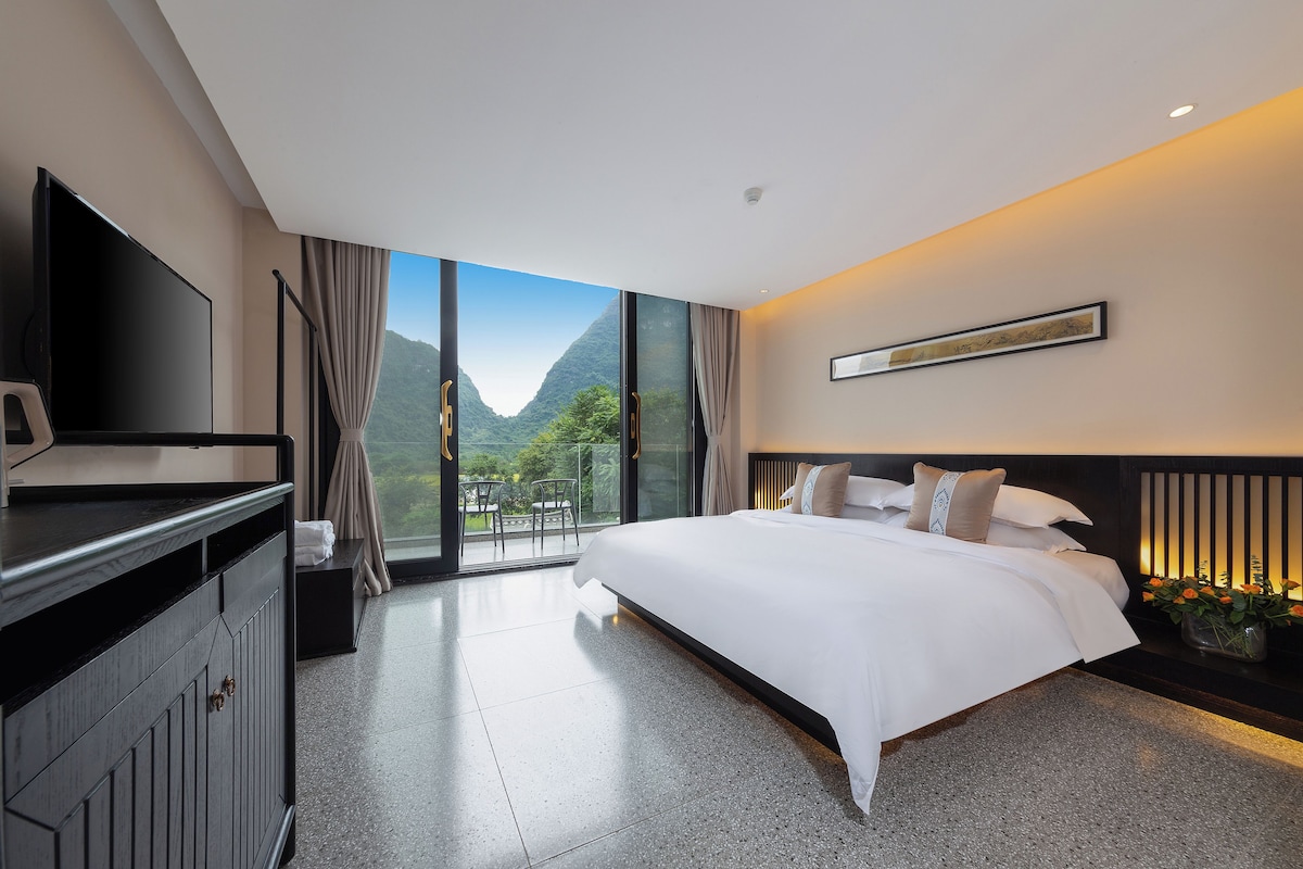 A modern hotel room with a large bed, a flat-screen TV, and a view of mountains through floor-to-ceiling windows.