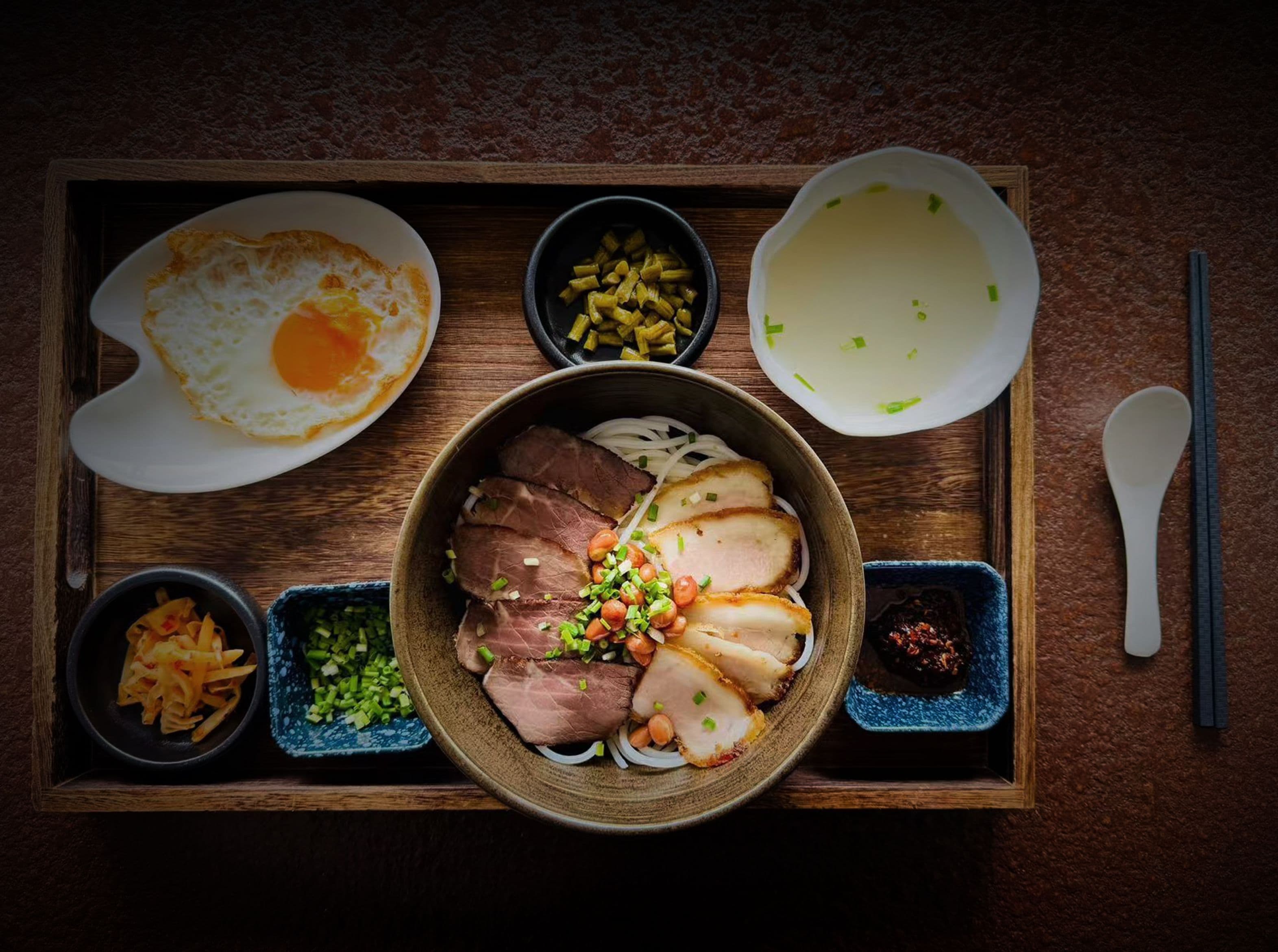 A meal tray with a bowl of sliced meat garnished with green onions and peanuts, a fried egg, and various side dishes including noodles and a bowl of soup.