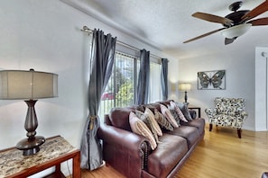 A living room with a leather sofa, two lamps, a butterfly wall art, and a ceiling fan. - Splash House at Indian Ridge | 4BR/2BA Kissimmee | Pool, Game Room (Kissimmee)
