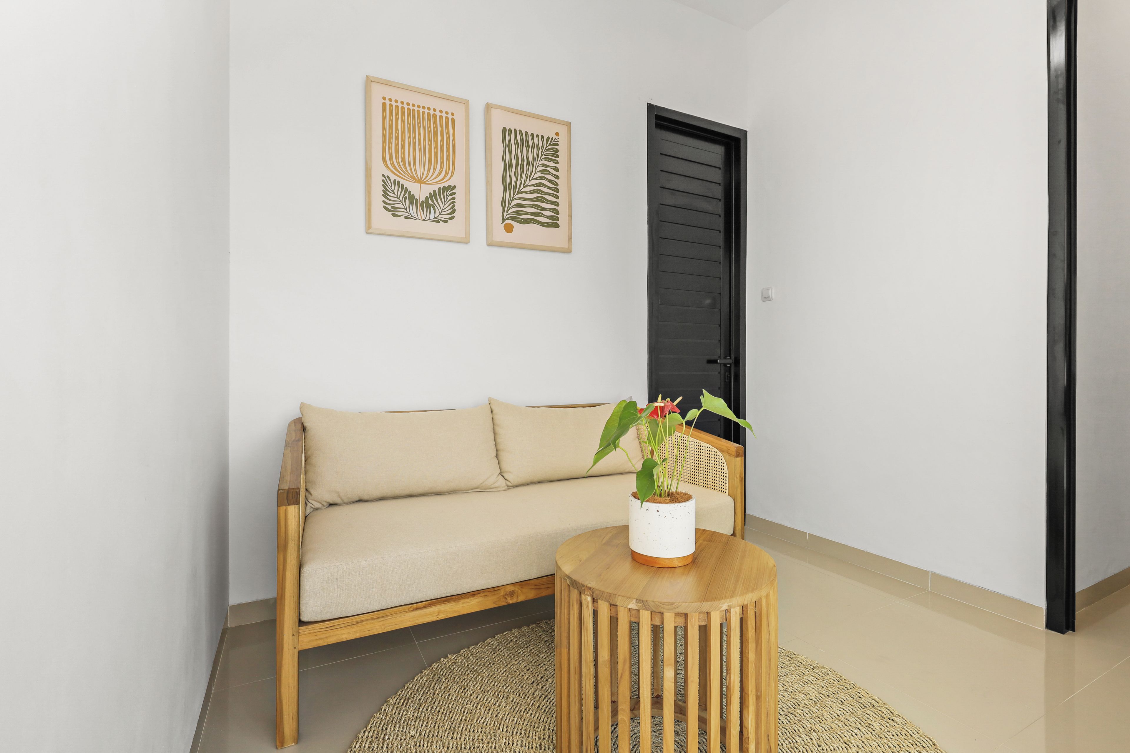 Een kamer met een beige bank, een ronde houten tafel met een plant erop, en twee ingelijste kunstwerken aan de muur.