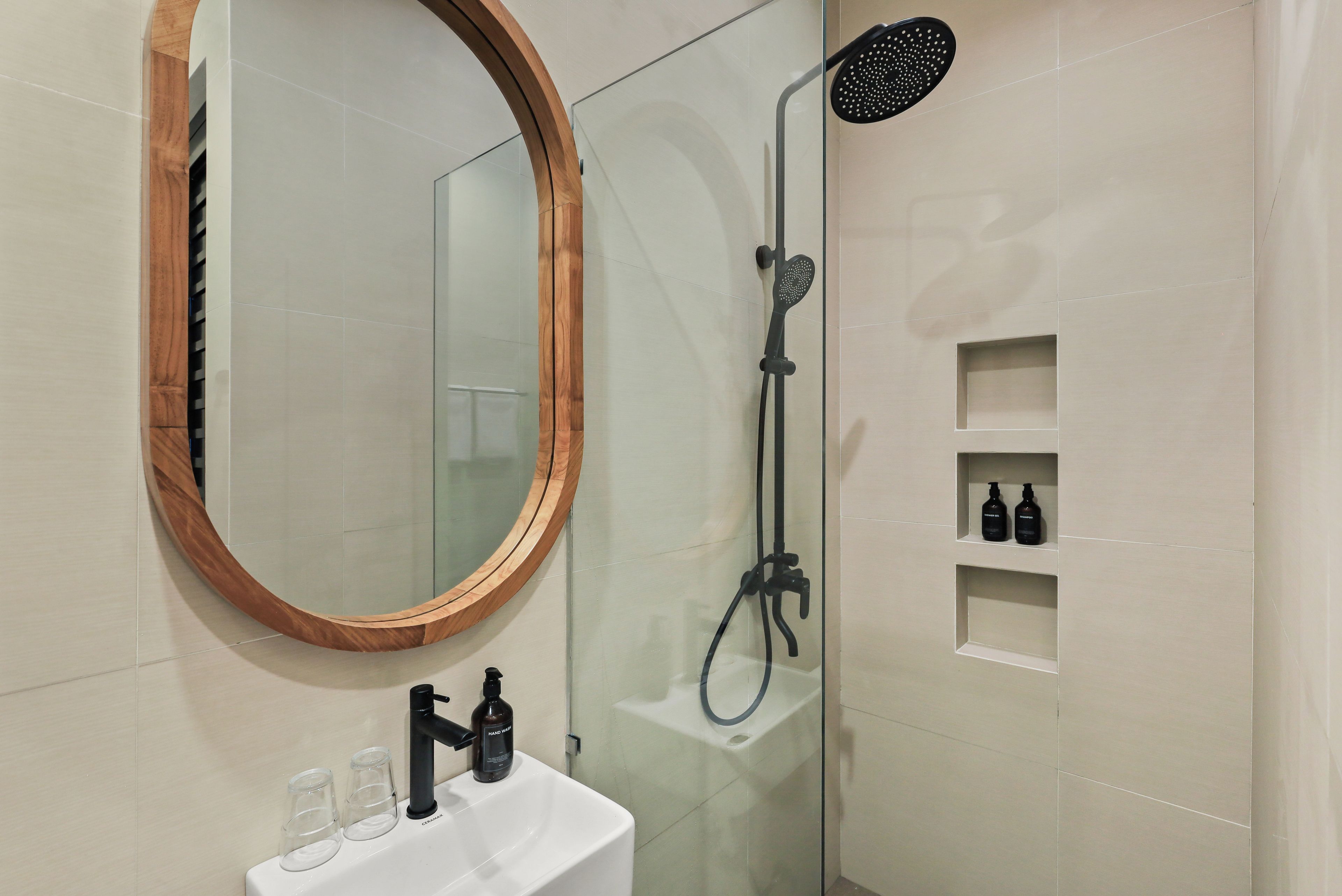 Une salle de bain avec un miroir encadré en bois, une douche avec un pommeau réglable, un lavabo blanc et deux flacons de savon pour les mains.