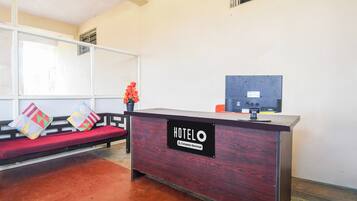 A hotel reception desk with a sign indicating it's company-serviced. There is a sofa with colorful cushions and a potted plant.