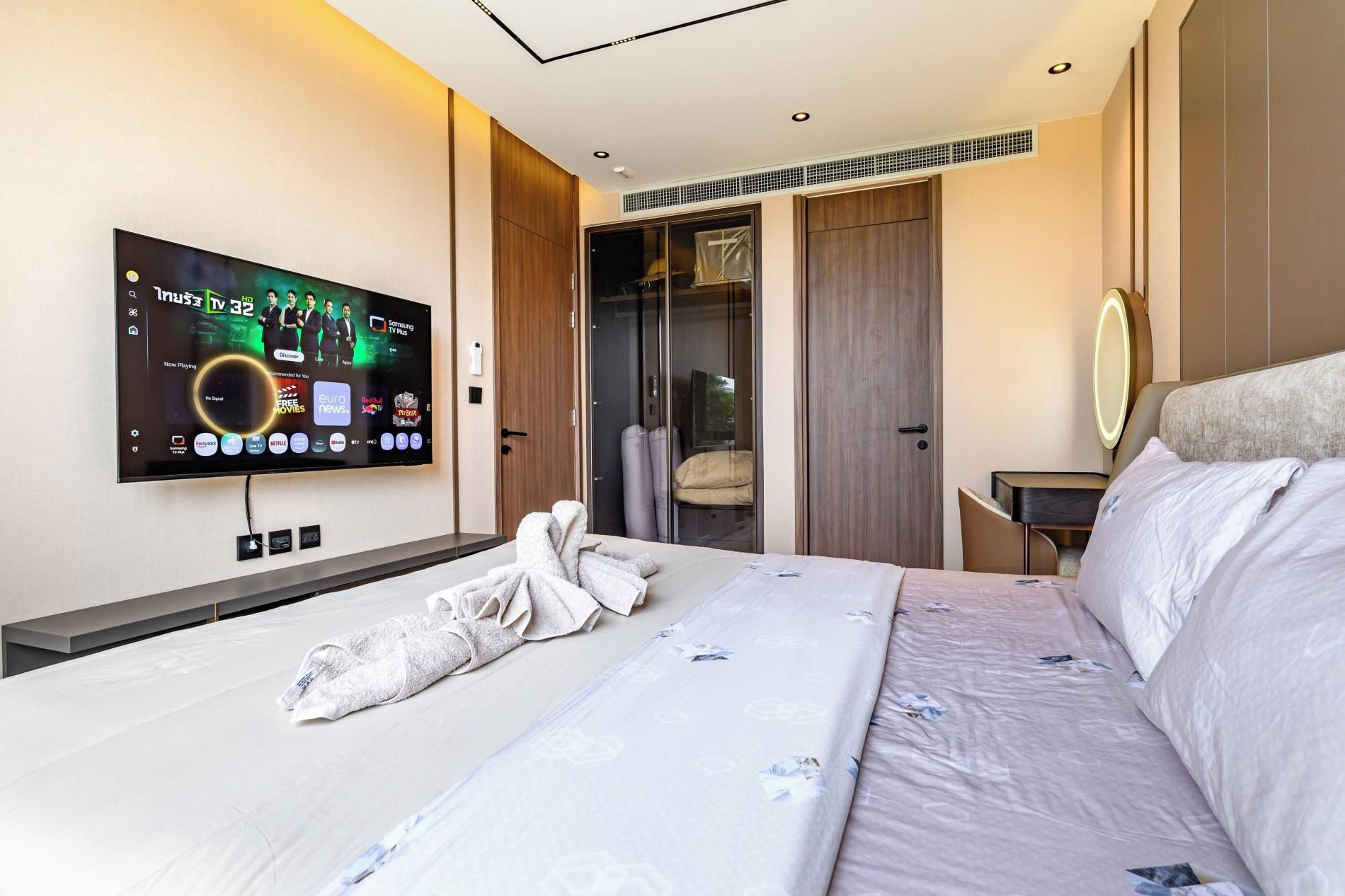Habitación de hotel con un televisor de pantalla plana grande montado en la pared, una cama con cubrecama blanco y estampado floral, una mesita de noche con lámpara y un espejo.