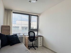 Ein Schlafzimmer mit einem Bett, einem Schreibtisch und einem Stuhl. Ein Fenster mit Jalousien ist zu sehen, und unter dem Schreibtisch ist eine Heizung erkennbar.