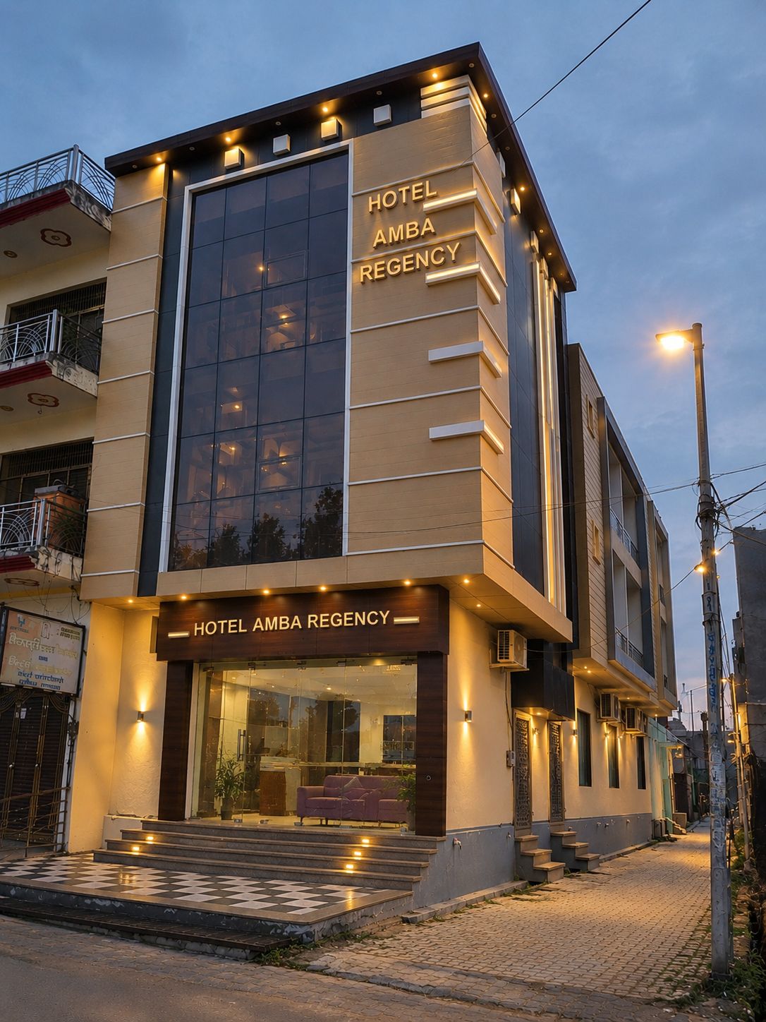 A modern hotel with a large glass facade, a prominent entrance with steps, and a sign indicating the name 'Hotel Amba Regency'.