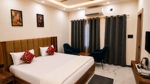 Una habitación de hotel con una cama grande, dos sillas, un escritorio, un equipo de aire acondicionado y obras de arte en las paredes.