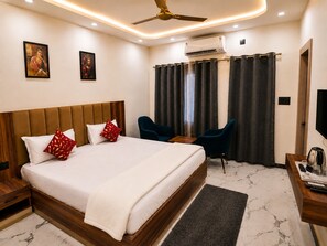A hotel room with a large bed, two chairs, a desk, an air conditioning unit, and artwork on the walls.