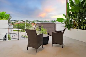 A balcony with wicker chairs and a round table, overlooking a residential area with houses and greenery. - Blanc Haven Canggu by Kerta Living (Badung)