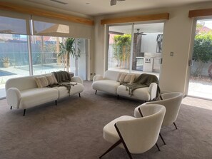 Ein modernes Wohnzimmer mit einer beigefarbenen Sofagarnitur, zwei Sesseln und einer großen Glasschiebetür zum Poolbereich.