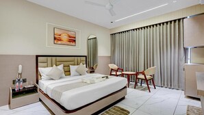 Habitación de hotel con una cama grande, una mesita de noche con teléfono y gafas, una silla, una pequeña mesa y un ventilador de techo.